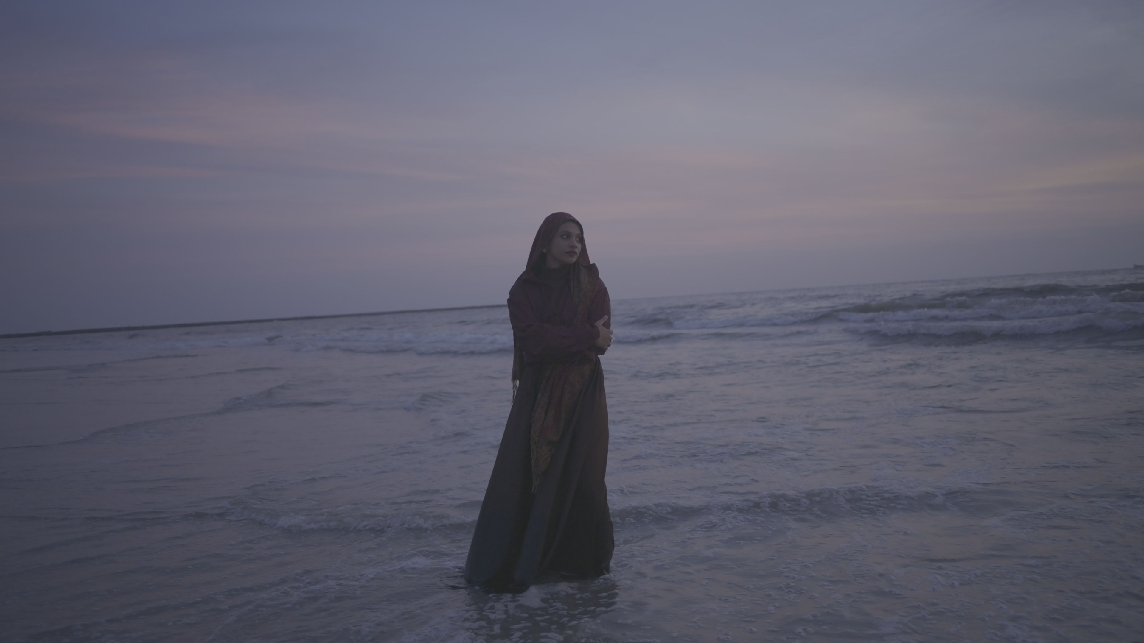 A woman in a dark robe and patterned shawl stands in the sea at dusk, as the camera recorded it in Log: flat, grey and low in contrast.