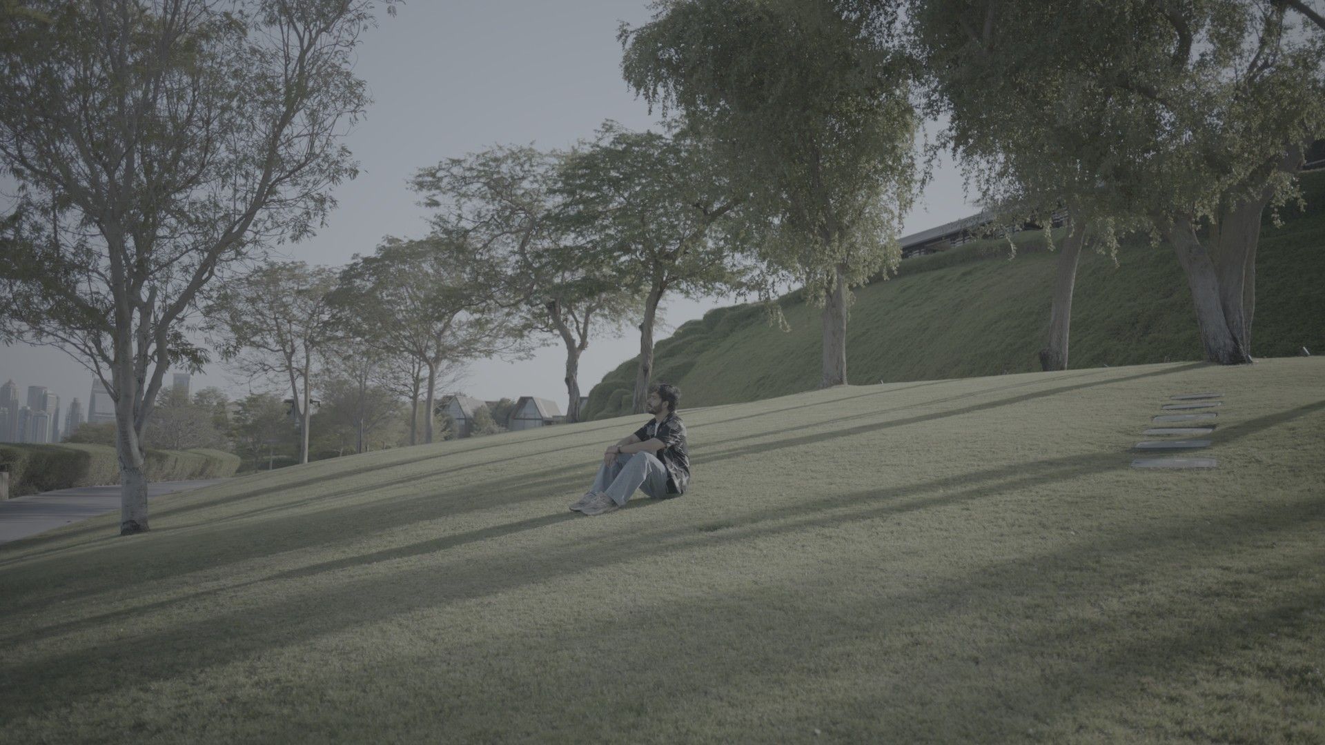 A man sits on a sloping lawn under trees in a park, as recorded in Log: flat, grey and washed out.